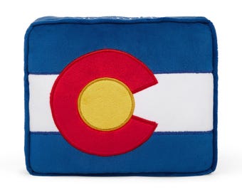 Pode incluir: Uma almofada de pelúcia azul, branca e vermelha em forma da bandeira do estado do Colorado. A bandeira apresenta um círculo vermelho grande com um círculo amarelo dentro e a letra "C" no centro.