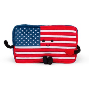 May include: A plush toy shaped like the American flag, featuring a blue corner with white stars and red and white stripes. The toy has a smiling face, black arms and feet, and a red heart. It is a patriotic and whimsical item.