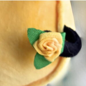 Puede incluir: Primer plano de un art&iacute;culo de tela amarilla con una rosa de fieltro y hojas verdes. La rosa es de un amarillo suave con un centro oscuro. Las hojas son verdes y hay un acento negro.
