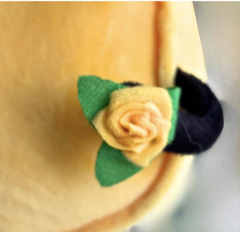Puede incluir: Primer plano de un art&iacute;culo de tela amarilla con una rosa de fieltro y hojas verdes. La rosa es de un amarillo suave con un centro oscuro. Las hojas son verdes y hay un acento negro.