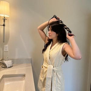 May include: A person in a white robe uses a curling iron to style their hair.