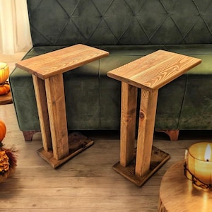 Może przedstawiać: Dwa drewniane stoliki pomocnicze z naturalnym wykończeniem drewna. Stoliki mają prosty design z prostokątnym blatem i dwoma pionowymi podporami. Stoliki znajdują się przed zieloną sofą.