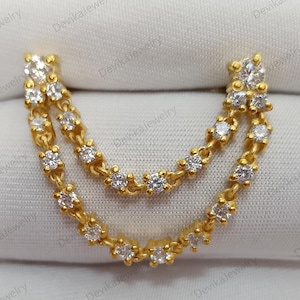 May include: A gold-colored earring with a double chain design. The earring features small, sparkling white gemstones set along the chains and at the top. The jewelry is displayed on a white surface.