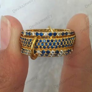 May include: A stackable gold ring set with blue and white gemstones. The rings are held together by a gold connector. The rings are held by a person's fingers. The brand name "DevikaJewelry" is visible in the background.
