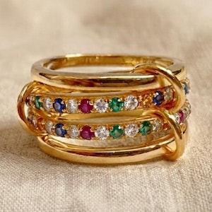 May include: A gold multi-band ring featuring two central bands embedded with alternating round-cut blue, white, red, and green gemstones. The design consists of several interlocking polished gold loops that create a layered, dimensional appearance.