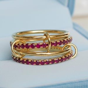 Puede incluir: Un anillo dorado con tres bandas, una con una fila de pequeñas piedras preciosas rojas. Las bandas están conectadas con pequeños aros dorados. El anillo se exhibe en una caja de joyería azul claro.