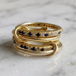 Può includere: Un set di anelli d'oro con un design unico e intrecciato. Ogni anello è adornato con piccole pietre preziose rotonde, blu scuro e chiare alternate. Gli anelli sono esposti su una superficie di marmo bianco.