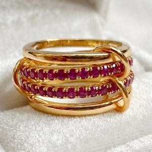 May include: A gold ring featuring a multi-band design with two central bands encrusted with small, round, vibrant red rubies. The structure consists of smooth, polished gold bands that interlock, creating a layered appearance with gemstone accents.