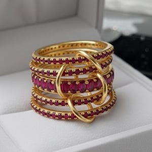 Op de afbeelding: Een stapel goudkleurige ringen met robijnkleurige edelstenen. De ringen zijn gerangschikt in een gelaagd ontwerp, met de edelstenen in een doorlopende band rond elke ring. De ringen zijn verbonden door goudkleurige lussen.