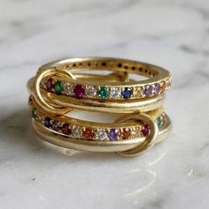 Op de afbeelding: Een gouden ring bestaande uit twee in elkaar grijpende banden. De ene band is van glad gepolijst goud, terwijl de andere is bezet met een rij kleine, veelkleurige edelstenen in rood, groen, blauw en wit, gerangschikt in een cirkelvormig patroon.