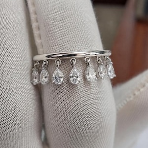 May include: A silver-toned ring featuring a thin, smooth band adorned with a series of seven dangling, pear-shaped clear stones. Each stone is individually suspended from the band by a small metal loop, creating a fringe-like effect.