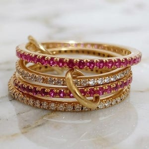 Op de afbeelding: Een stapel goudkleurige ringen. Sommige ringen zijn versierd met kleine, levendige roze edelstenen, terwijl andere heldere, sprankelende stenen hebben. De ringen zijn in elkaar verweven, waardoor een uniek en opvallend ontwerp ontstaat.