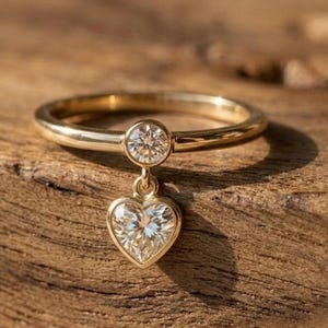 Może przedstawiać: Złoty pierścionek z okrągłym diamentem i zawieszką w kształcie serca z diamentami. Pierścionek leży na drewnianej powierzchni, a światło słoneczne odbija się w diamentach. To biżuteria.