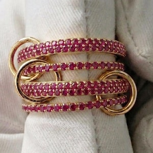 Puede incluir: Anillo dorado con cuatro bandas unidas, cada una incrustada con pequeñas y vibrantes piedras rojas. El diseño del anillo presenta bucles entrelazados, creando una pieza audaz. Las piedras están muy juntas.