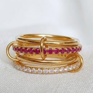 Può includere: Un anello dorato a più fasce composto da quattro anelli distinti interconnessi. Una fascia è incastonata con piccole pietre rosse vivaci, un'altra con pietre chiare scintillanti e due fasce dorate lucide, collegate da una barra verticale.