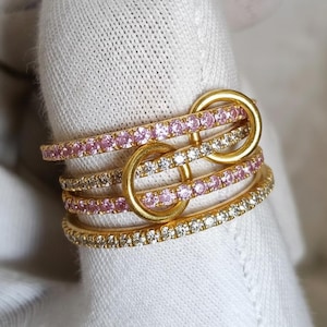 Puede incluir: Un anillo de oro con múltiples bandas, adornado con piedras preciosas rosas y transparentes. Las bandas están unidas con aros dorados. El anillo está sobre un fondo blanco.