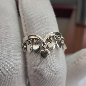 May include: A silver ring with a heart-shaped design. The ring features a series of small, dangling hearts suspended from the band. The ring is shown on a white surface.