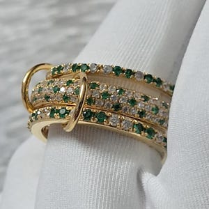 Op de afbeelding: Een goudkleurige ring met meerdere banden, elk bezet met kleine groene en heldere edelstenen. Het ontwerp van de ring is ingewikkeld, waarbij de edelstenen een sprankelend effect creëren. Gepresenteerd tegen een witte stof, die de kleuren goud en edelstenen benadrukt.