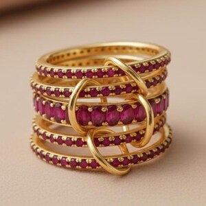K&ouml;nnte beinhalten: Ein Stapel goldfarbener Ringe mit einem Band aus leuchtend rubinroten Edelsteinen. Die Ringe sind mit goldenen B&ouml;gen verbunden, wodurch ein einzigartiges Design entsteht. Der Schmuck wird auf einem neutralen Hintergrund pr&auml;sentiert.