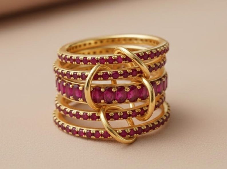 K&ouml;nnte beinhalten: Ein Stapel goldfarbener Ringe mit einem Band aus leuchtend rubinroten Edelsteinen. Die Ringe sind mit goldenen B&ouml;gen verbunden, wodurch ein einzigartiges Design entsteht. Der Schmuck wird auf einem neutralen Hintergrund pr&auml;sentiert.