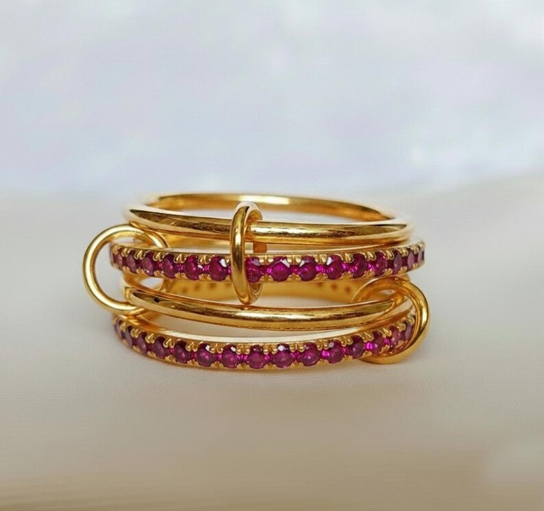 Op de afbeelding: Een stapelbare set gouden ringen, verbonden door gouden lussen en een centrale ring. Elke ring is versierd met kleine, rode edelstenen, wat een luxueus ontwerp cre&euml;ert. De ringen staan tegen een zachte, neutrale achtergrond.