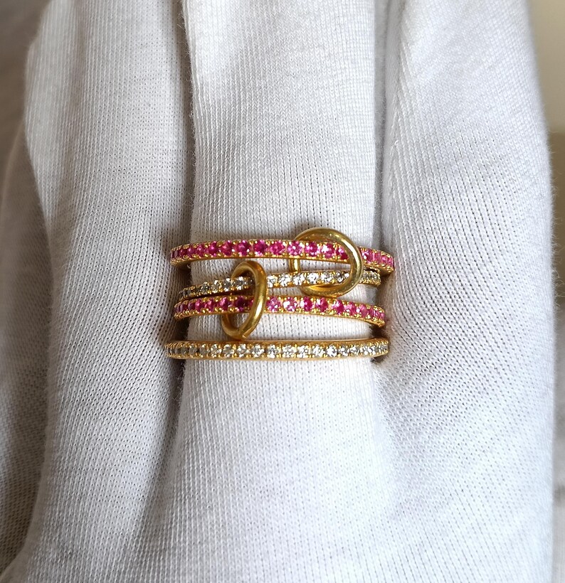 Puede incluir: Un conjunto de anillos de oro con varios dise&ntilde;os. Algunos anillos est&aacute;n adornados con piedras preciosas rosas, mientras que otros presentan piedras preciosas claras. Los anillos est&aacute;n entrelazados con bucles dorados, creando un aspecto &uacute;nico y elegante.