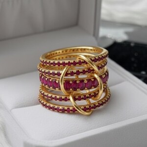 K&ouml;nnte beinhalten: Ein Stapel goldfarbener Ringe mit rubinroten Edelsteinen. Die Ringe sind in einem geschichteten Design angeordnet, wobei die Edelsteine in einem durchgehenden Band um jeden Ring gesetzt sind. Die Ringe sind durch goldfarbene Schlaufen verbunden.