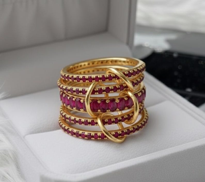 K&ouml;nnte beinhalten: Ein Stapel goldfarbener Ringe mit rubinroten Edelsteinen. Die Ringe sind in einem geschichteten Design angeordnet, wobei die Edelsteine in einem durchgehenden Band um jeden Ring gesetzt sind. Die Ringe sind durch goldfarbene Schlaufen verbunden.
