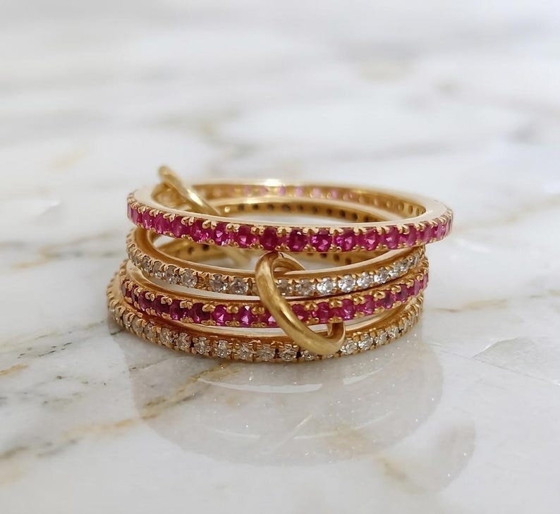 Puede incluir: Una pila de anillos dorados. Algunos anillos est&aacute;n adornados con peque&ntilde;as y vibrantes piedras preciosas rosas, mientras que otros presentan piedras claras y brillantes. Los anillos est&aacute;n entrelazados, creando un dise&ntilde;o &uacute;nico y llamativo.