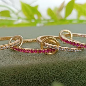 Puede incluir: Cuatro anillos de oro entrelazados, cada uno con un dise&ntilde;o diferente. Dos anillos est&aacute;n incrustados con peque&ntilde;as piedras claras y dos est&aacute;n adornados con peque&ntilde;as piedras rosas. Los anillos se exhiben sobre una superficie verde.