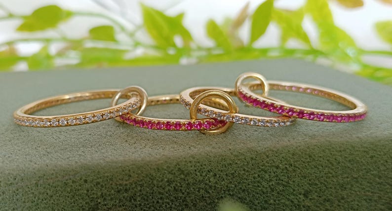 Puede incluir: Cuatro anillos de oro entrelazados, cada uno con un dise&ntilde;o diferente. Dos anillos est&aacute;n incrustados con peque&ntilde;as piedras claras y dos est&aacute;n adornados con peque&ntilde;as piedras rosas. Los anillos se exhiben sobre una superficie verde.