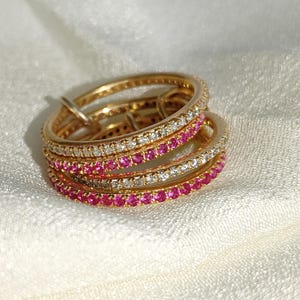 Puede incluir: Un conjunto de anillos dorados con filas alternas de piedras preciosas blancas y rosas. Los anillos est&aacute;n colocados sobre una superficie blanca y texturizada. Las piedras preciosas son peque&ntilde;as y est&aacute;n espaciadas uniformemente.