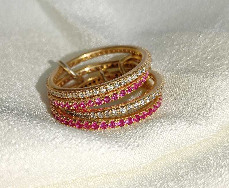 Puede incluir: Un conjunto de anillos dorados con filas alternas de piedras preciosas blancas y rosas. Los anillos est&aacute;n colocados sobre una superficie blanca y texturizada. Las piedras preciosas son peque&ntilde;as y est&aacute;n espaciadas uniformemente.