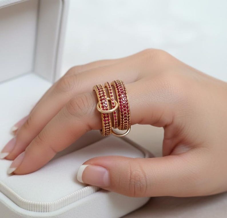 May include: A gold ring featuring three bands, each set with small, red gemstones. The bands are intertwined, forming a distinctive design. The ring is shown on a finger, highlighting its intricate details and vivid colour. The ring's design is eye-catching.