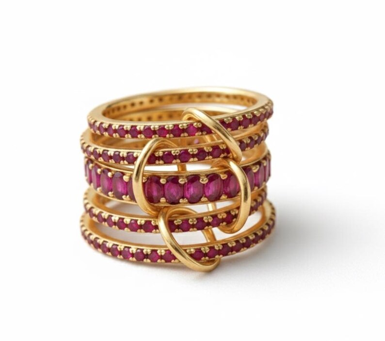 K&ouml;nnte beinhalten: Ein Stapel goldener Ringe mit rubinroten Edelsteinen. Die Ringe sind durch goldene Schlaufen verbunden, wodurch ein einzigartiges und auff&auml;lliges Design entsteht. Die Edelsteine sind eng gesetzt, auf wei&szlig;em Hintergrund.