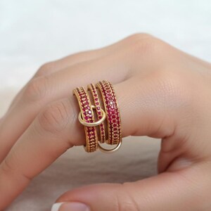 May include: A gold ring featuring three stacked bands, each set with small, red gemstones. A smaller, gold ring connects the bands. The ring is worn on a finger.