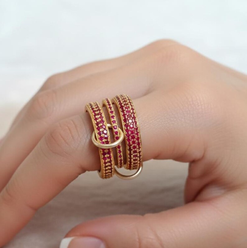 May include: A gold ring featuring three stacked bands, each set with small, red gemstones. A smaller, gold ring connects the bands. The ring is worn on a finger.