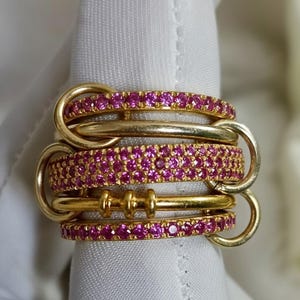 May include: A gold ring set with multiple bands. Two bands are set with small, vibrant pink gemstones. The bands are connected by gold loops and bars, creating a unique, layered design. The ring is displayed on a white surface.