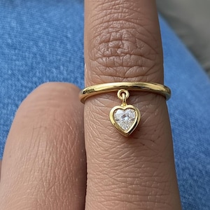 Puede incluir: Un anillo de oro con un colgante en forma de corazón. El colgante es un diamante en forma de corazón engastado en un bisel de oro. El anillo está en un dedo, sobre un fondo de mezclilla azul.