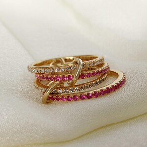 Puede incluir: Un conjunto de anillos dorados con piedras preciosas incrustadas. Dos anillos est&aacute;n adornados con peque&ntilde;as piedras transparentes, mientras que otros dos presentan vibrantes piedras rosas. Los anillos est&aacute;n entrelazados y descansan sobre una superficie blanca y sedosa.