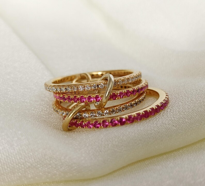 Puede incluir: Un conjunto de anillos dorados con piedras preciosas incrustadas. Dos anillos est&aacute;n adornados con peque&ntilde;as piedras transparentes, mientras que otros dos presentan vibrantes piedras rosas. Los anillos est&aacute;n entrelazados y descansan sobre una superficie blanca y sedosa.