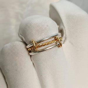 Può includere: Un anello in argento e oro. L'anello presenta una fascia dorata intrecciata tra due fasce argentate, con piccoli connettori dorati. L'anello è mostrato su una superficie bianca.