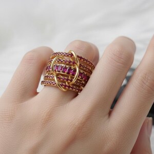 K&ouml;nnte beinhalten: Ein goldfarbener Ring mit mehreren B&auml;ndern, die jeweils mit kleinen, roten Edelsteinen besetzt sind. Der Ring hat ein einzigartiges Design mit einer schleifenf&ouml;rmigen, ineinander verschlungenen Struktur. Die Edelsteine sind tiefrot, und der Ring ist vor einem neutralen Hintergrund platziert.