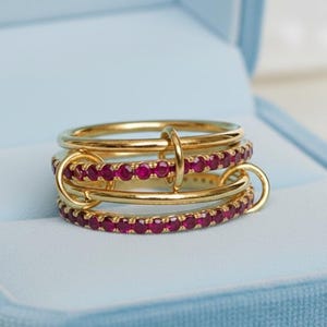 Op de afbeelding: Een goudkleurige ring met drie banden, waarvan &eacute;&eacute;n met een rij kleine, rode edelstenen. De banden zijn verbonden met kleine gouden ringen. De ring wordt gepresenteerd in een lichtblauwe sieradendoos.