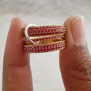 May include: Several gold-coloured rings, each set with numerous small, bright red gemstones. One silver-toned ring is interlocked with the others. The rings are held between fingers, highlighting the jewellery's design and colour.