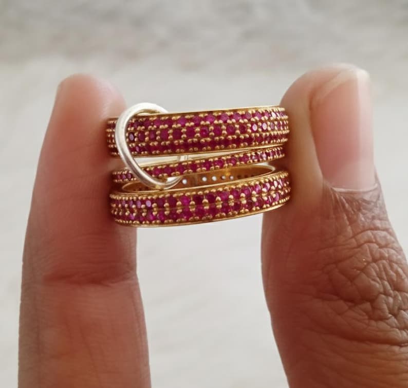 May include: Several gold-coloured rings, each set with numerous small, bright red gemstones. One silver-toned ring is interlocked with the others. The rings are held between fingers, highlighting the jewellery's design and colour.