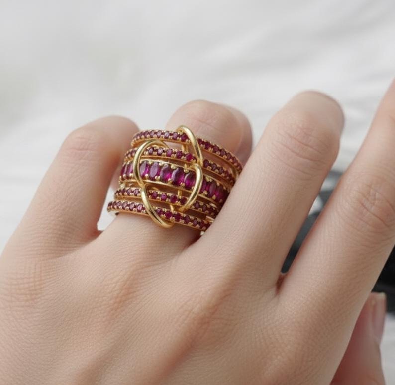 K&ouml;nnte beinhalten: Ein goldfarbener Ring mit mehreren B&auml;ndern, die jeweils mit kleinen, roten Edelsteinen besetzt sind. Der Ring hat ein einzigartiges Design mit einer schleifenf&ouml;rmigen, ineinander verschlungenen Struktur. Die Edelsteine sind tiefrot, und der Ring ist vor einem neutralen Hintergrund platziert.
