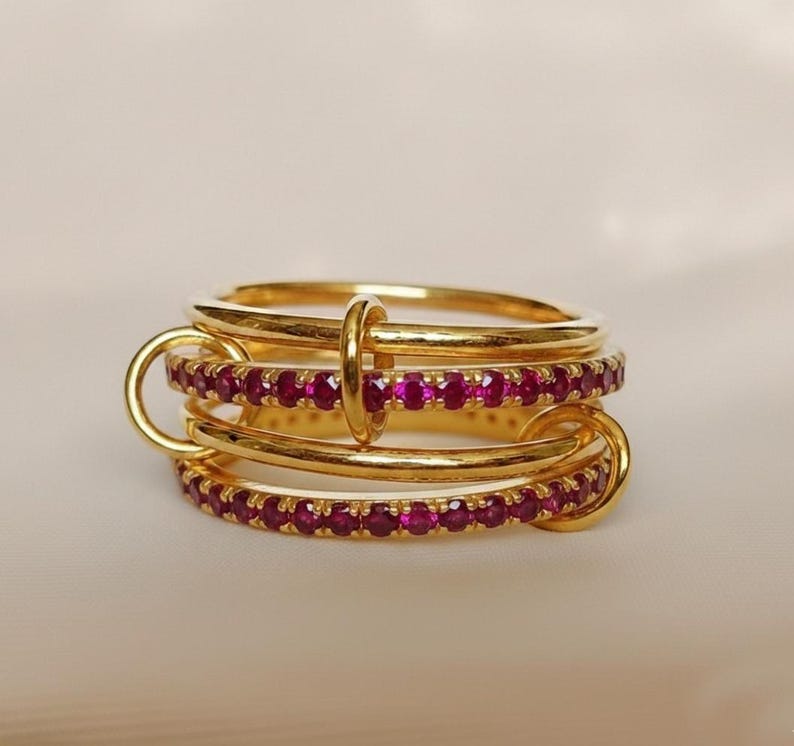 Op de afbeelding: Een goudkleurige ring met drie in elkaar grijpende banden. Twee banden zijn bezet met kleine, levendige rode edelstenen. De ring heeft een uniek ontwerp met verbindingslussen en een centrale verticale staaf.