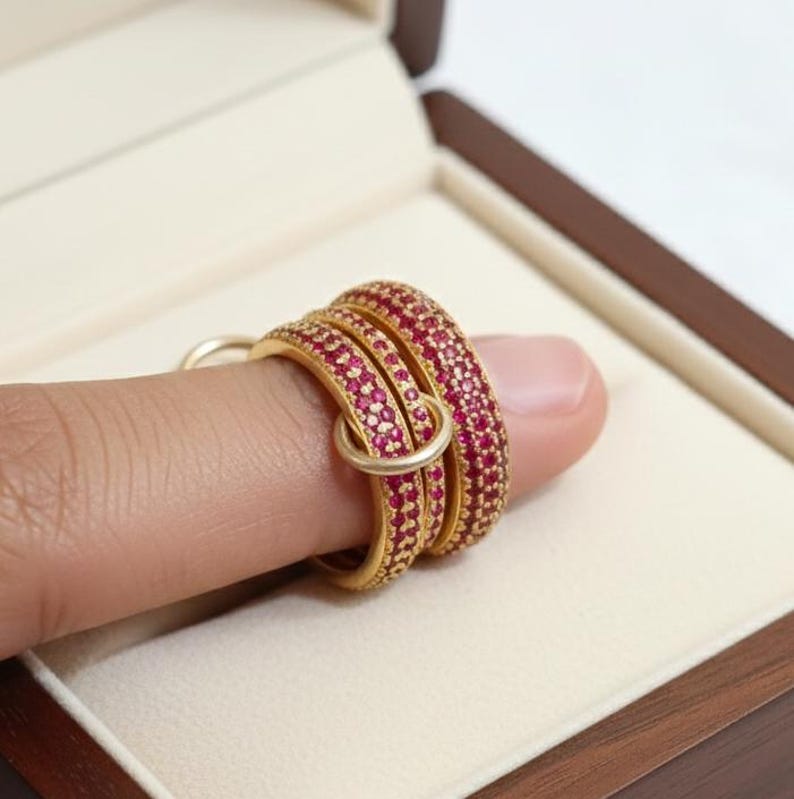 May include: A close-up of gold rings adorned with red gemstones. The rings are stacked on a finger, showcasing their intricate design. The small red gemstones are closely set, covering the surface of the rings. The rings are presented inside a brown jewellery box.