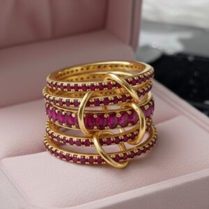 K&ouml;nnte beinhalten: Ein Stapel goldfarbener Ringe mit einem Band aus leuchtend rubinroten Edelsteinen. Die Ringe sind mit goldenen Schlaufen verbunden, wodurch ein einzigartiges Design entsteht. Der Schmuck wird in einer rosa Schmuckschatulle pr&auml;sentiert.